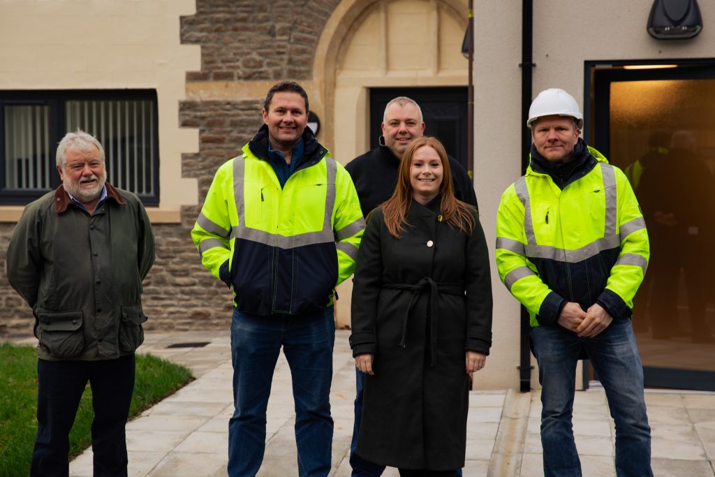 Trivallis Housing Landlord Wales Five people standing in front of a building. Two are wearing high-visibility jackets, and one is wearing a white hard hat. A woman in the center is wearing a black coat. The background shows a stone wall and window.