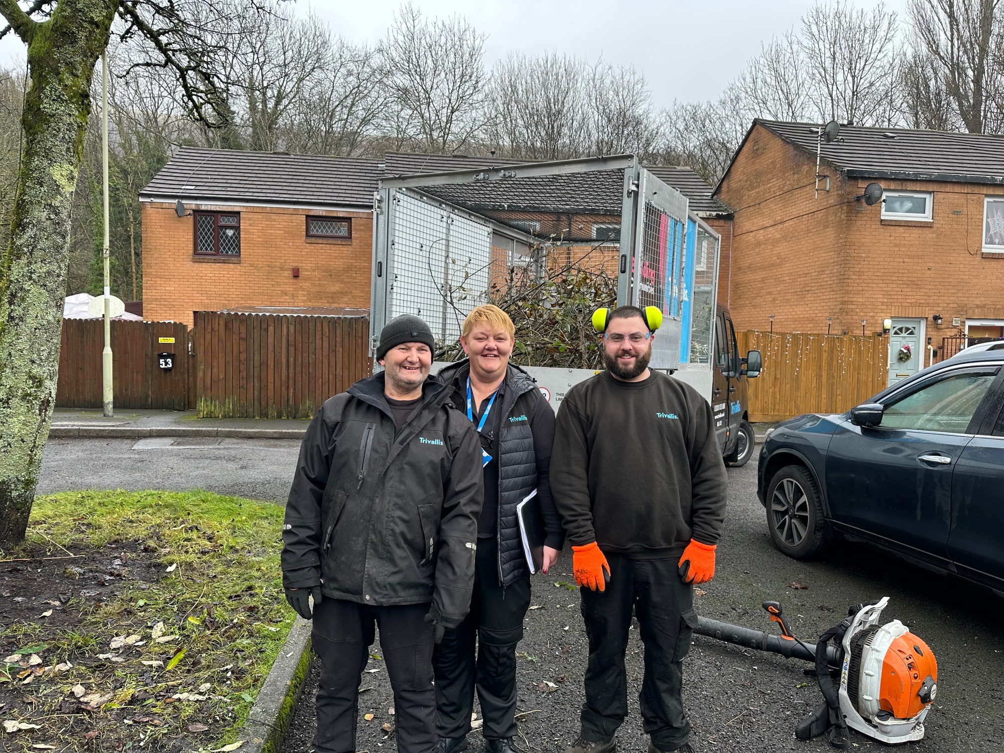 Trivallis Housing Landlord Wales Three people stand outdoors on a residential street, with a truck and some tools nearby. They are dressed in work clothing suitable for outdoor tasks. Two houses and some trees are visible in the background.