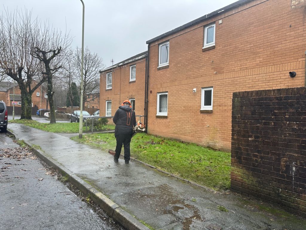 Trivallis Housing Landlord Wales A person in a dark jacket is using a leaf blower on a wet sidewalk next to a brick building. There are bare trees and a few parked vehicles in the background. The sky is overcast.