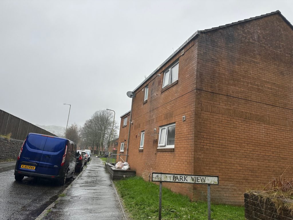 Trivallis Housing Landlord Wales A brick residential building is situated on a wet street with parked vehicles. A sign reading "Park View" is visible in the foreground. The sky is overcast, and trees line the street in the background.