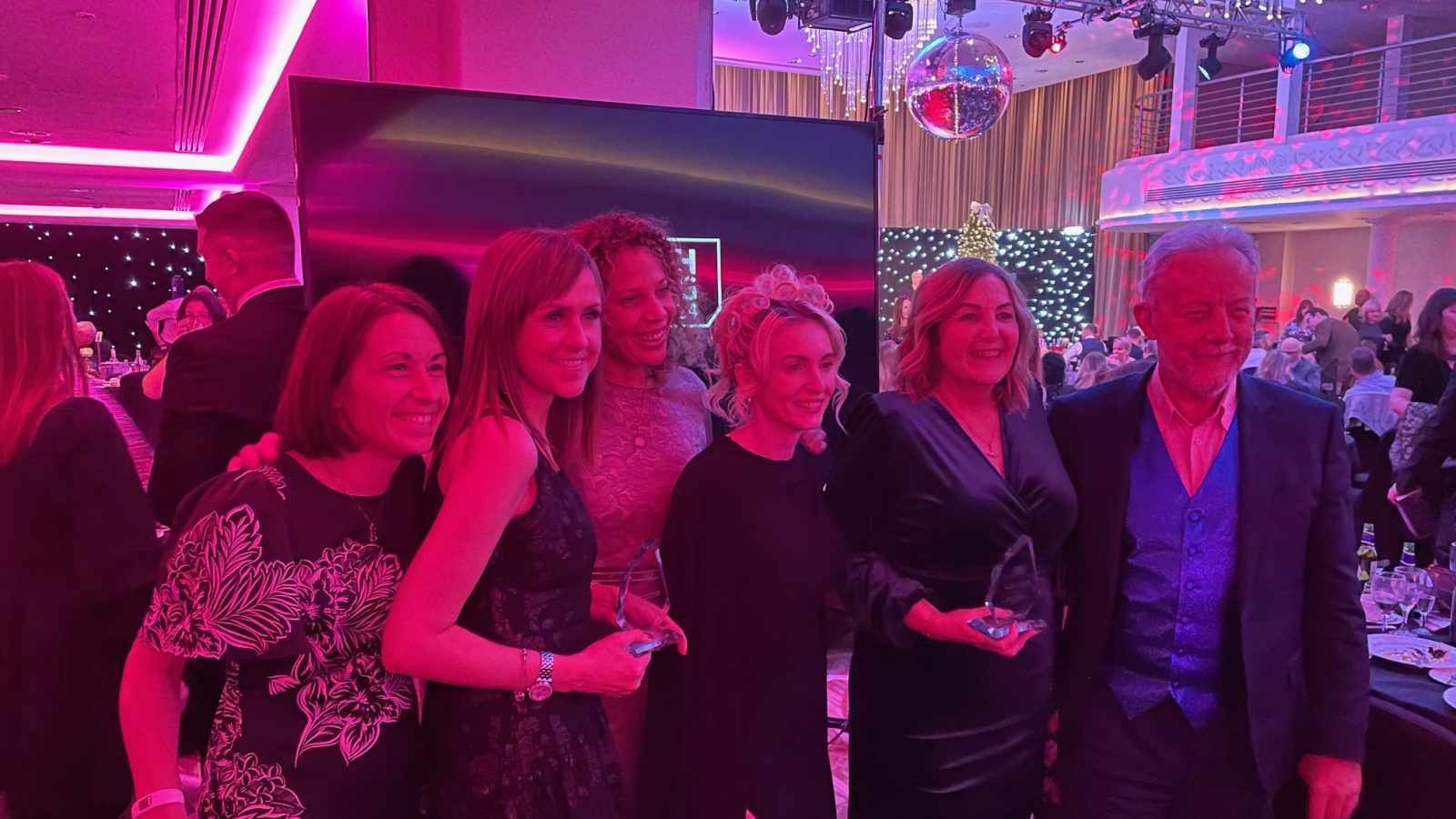 Trivallis Housing Landlord Wales A group of six people dressed in formal attire pose for a photo at an event with dim pink and purple lighting. They are standing in front of a starry backdrop inside a venue with chandeliers and a disco ball. Some are holding awards.