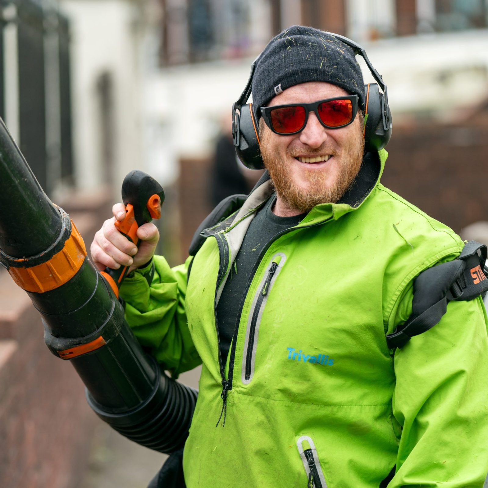 Trivallis Housing Landlord Wales A person wearing sunglasses, earmuffs, and a green jacket operates a leaf blower on a sidewalk. They are smiling and holding the blower's handle, with a fence and residential buildings visible in the background.