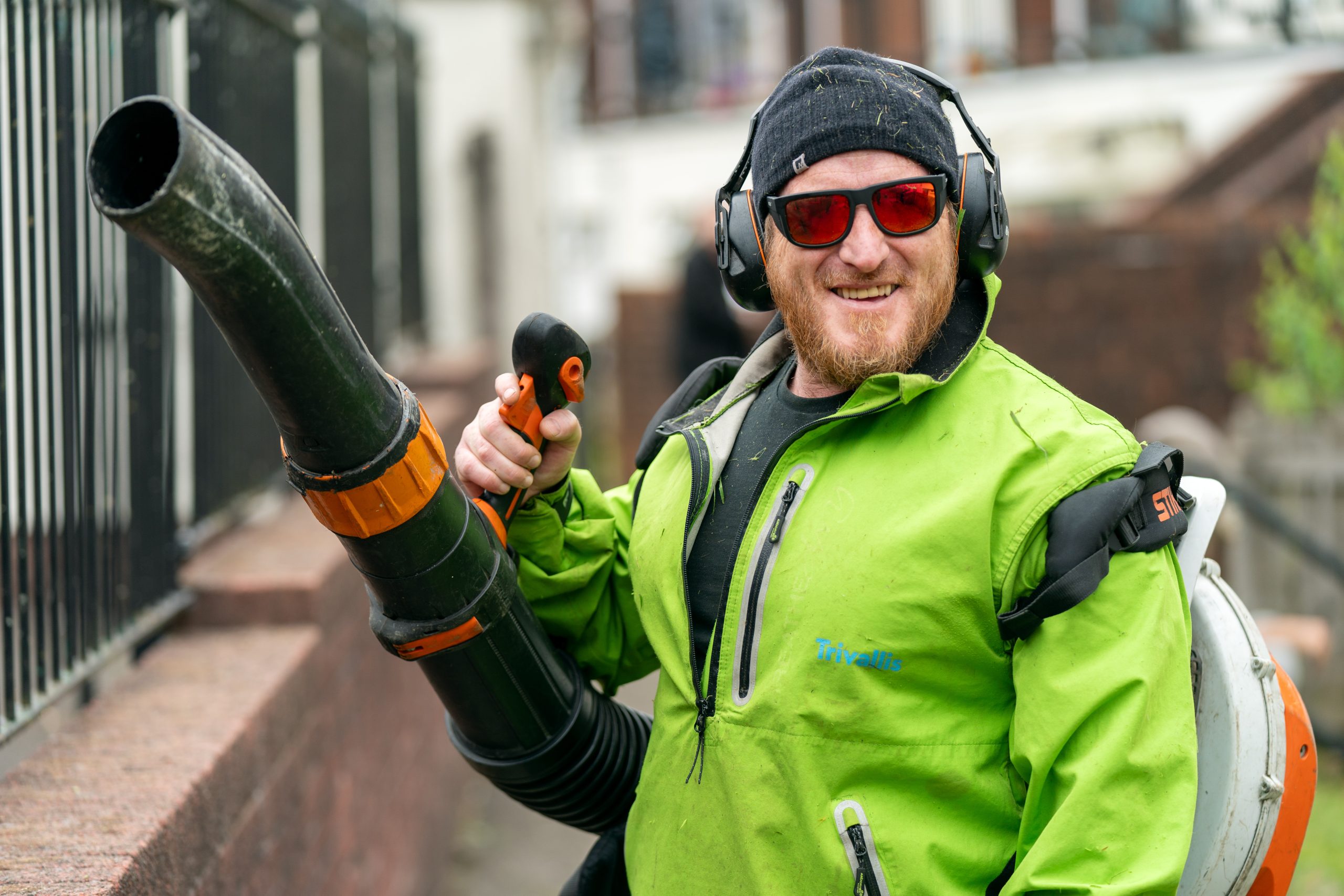 Trivallis Housing Landlord Wales A person wearing sunglasses, earmuffs, and a green jacket operates a leaf blower on a sidewalk. They are smiling and holding the blower's handle, with a fence and residential buildings visible in the background.