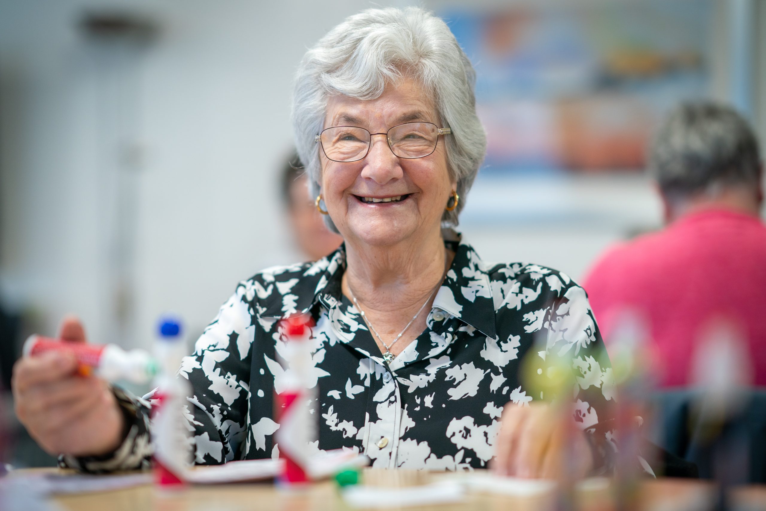 Trivallis Housing Landlord Wales An elderly woman with gray hair and glasses is smiling while holding bingo chips. She wears a black and white patterned blouse and is seated at a table. Other people are visible in the blurred background.