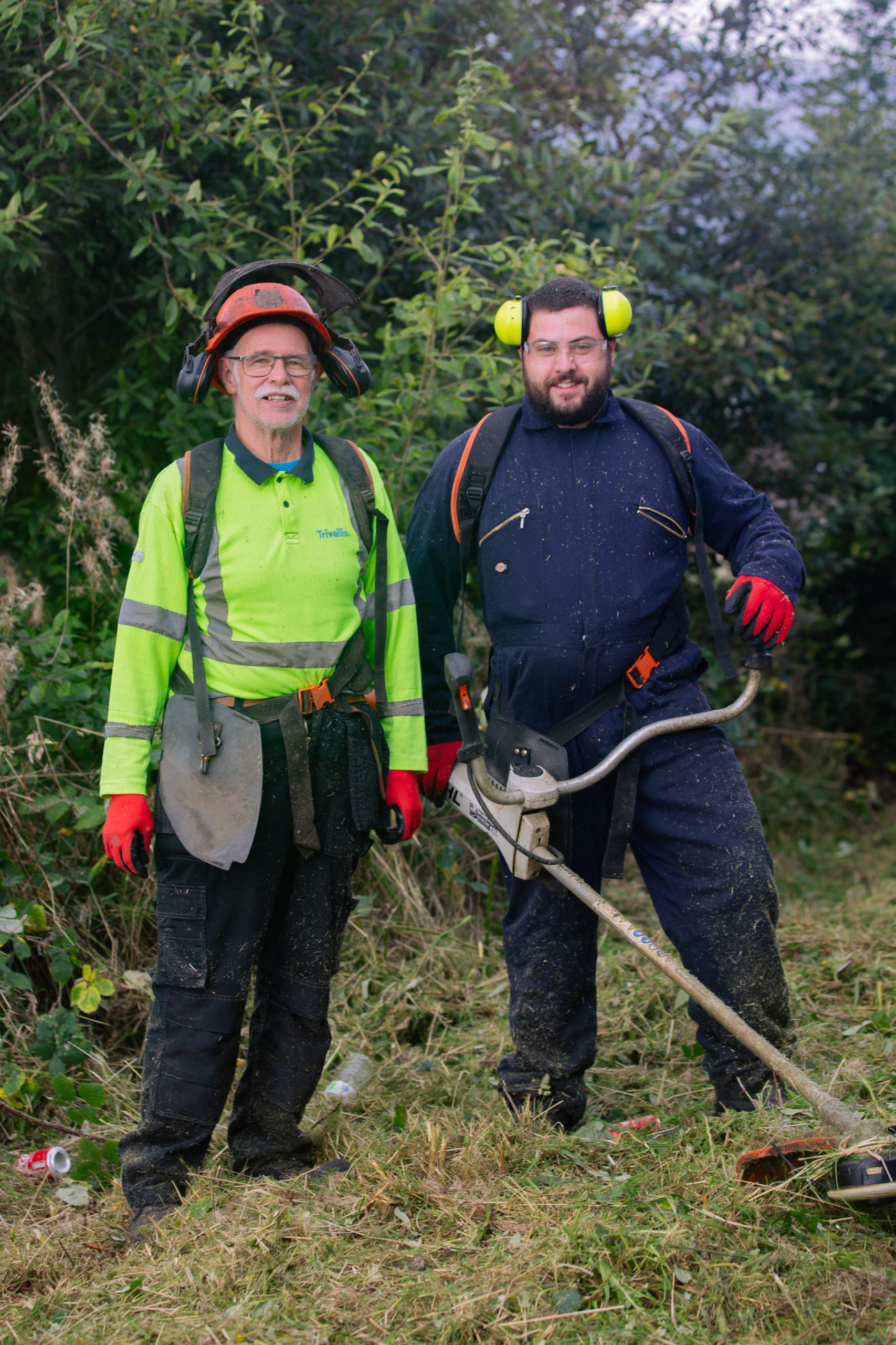 Trivallis Housing Landlord Wales Two people pose outdoors in work attire. One wears a green safety jacket and hard hat, the other a dark jumpsuit with ear protection. Both hold equipment for landscaping, standing on grass with foliage in the background.