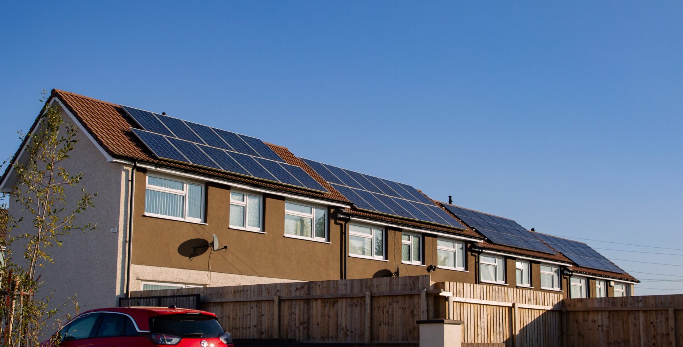 Trivallis Housing Landlord Wales A row of houses with solar panels on the roofs is shown under a clear blue sky. A red car is parked in the driveway next to a wooden fence. The scene is well lit by sunlight.