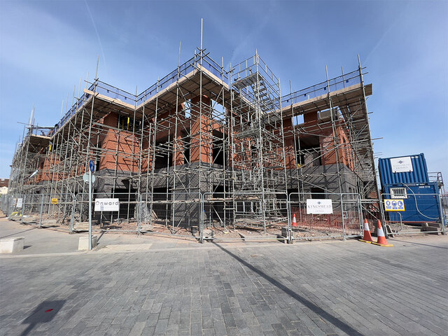 Trivallis Housing Landlord Wales A brick building under construction with surrounding metal scaffolding. The structure has two visible floors, and there are construction cones and a blue container nearby. The sky is clear and the ground is paved with gray bricks.