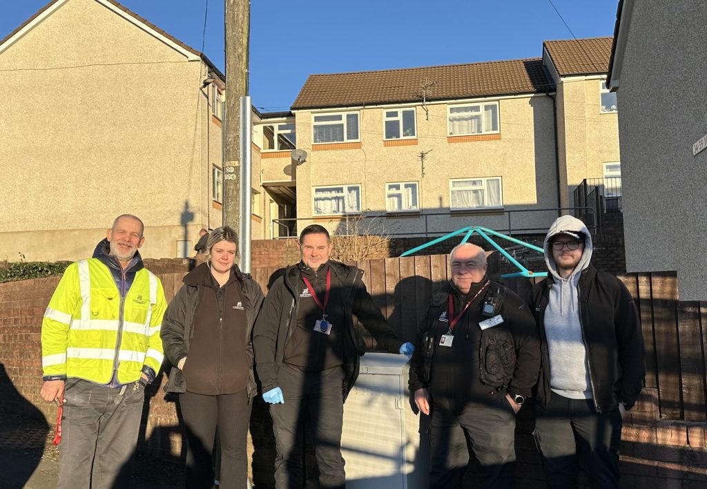 Trivallis Housing Landlord Wales Five people stand in front of a residential area with houses under a clear blue sky. One person wears a high-visibility jacket. A white van is partially visible on the left. They are standing on a street with a wooden fence behind them.