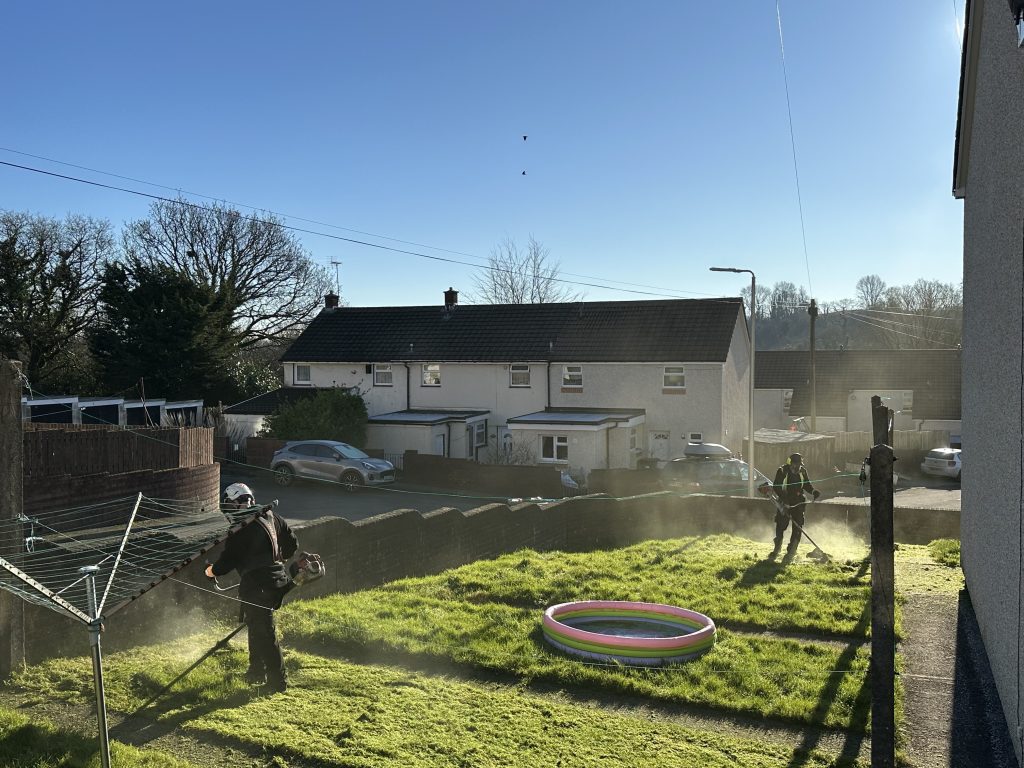 Trivallis Housing Landlord Wales Two people are using grass trimmers on a sunny day in a suburban backyard. The area has a small inflatable pool and drying rack. A house is visible in the background with a car parked nearby. The sky is clear and blue.