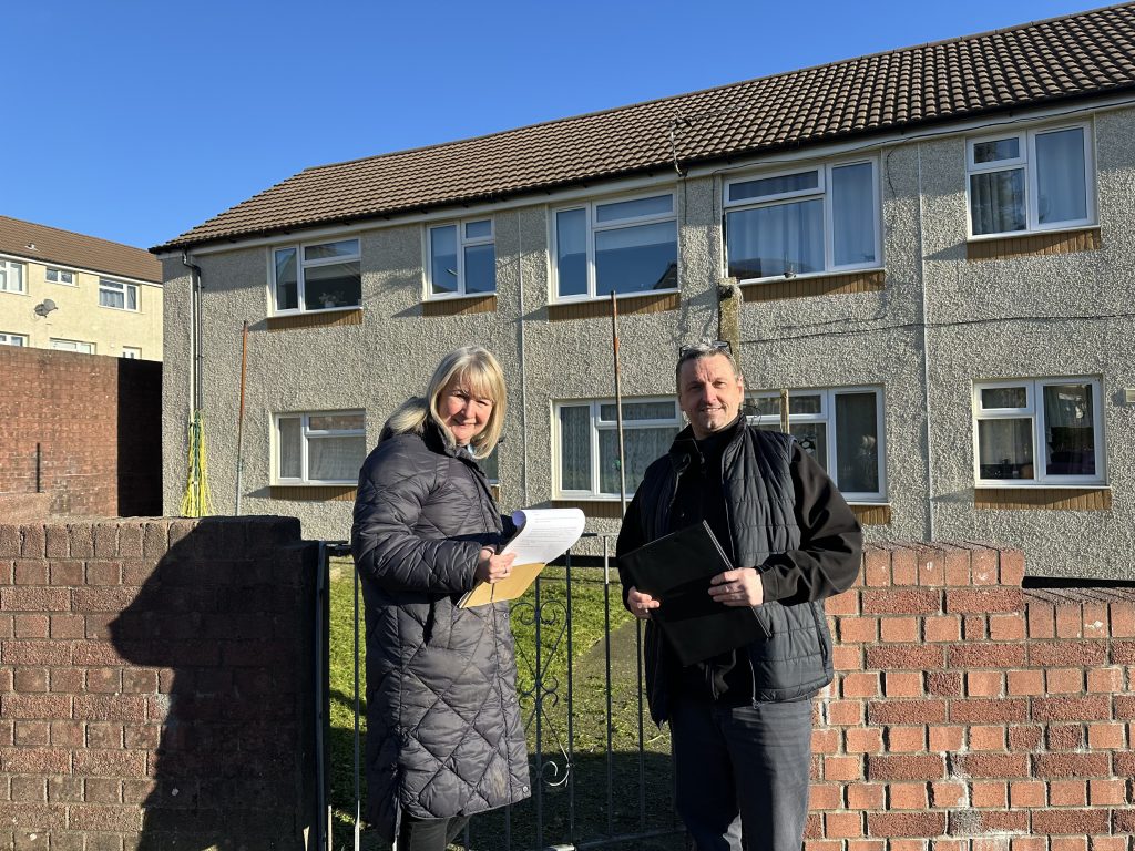 Trivallis Housing Landlord Wales A woman in a black coat and a man in a dark jacket stand in front of a two-story building. They hold papers and a tablet. A brick wall and metal gate are in the foreground. It is a clear, sunny day.