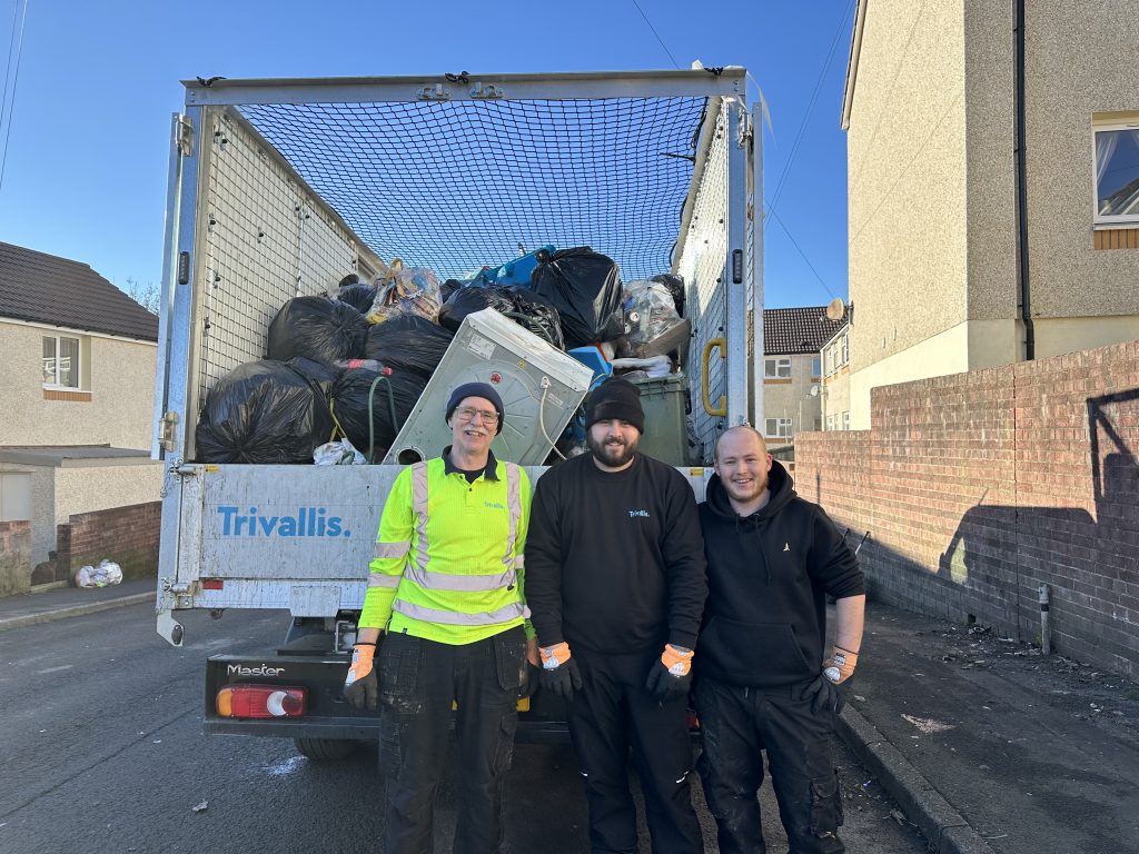 Trivallis Housing Landlord Wales Three people in work attire stand smiling in front of a Trivallis truck filled with black garbage bags and assorted items. The scene is on a residential street with houses visible in the background.