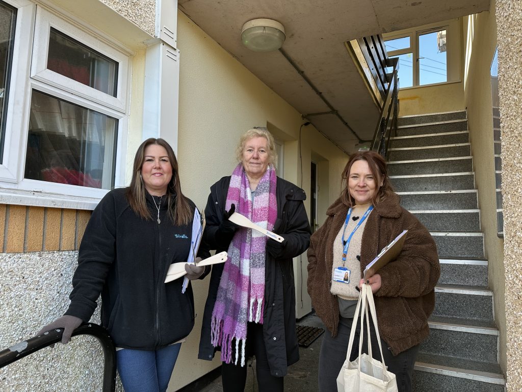 Trivallis Housing Landlord Wales Three women stand in front of a staircase in a residential area. They are dressed warmly, holding papers and a tote bag. The building has a beige exterior with a white window. The staircase is on the right side of the image.