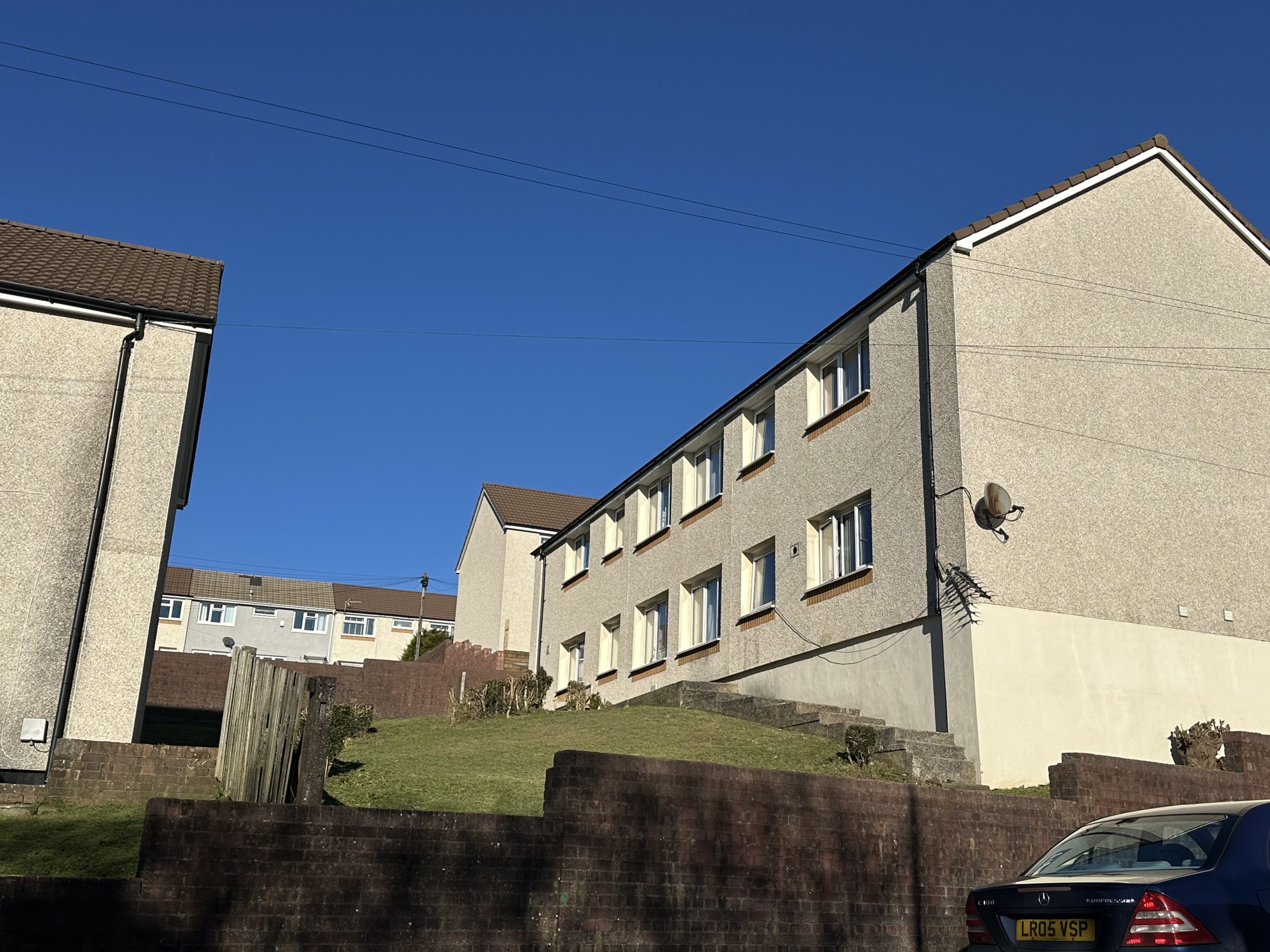 Trivallis Housing Landlord Wales A two-story apartment building with a white exterior is situated on a sloped grassy hill. The building has several windows and a satellite dish. Nearby are additional buildings and a car parked in the foreground. The sky is clear and blue.