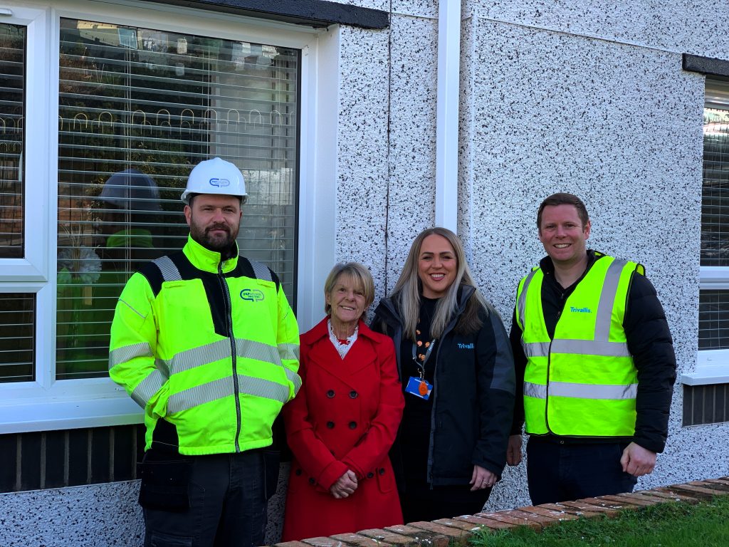 Trivallis Housing Landlord Wales Four people are standing in front of a building with a window. Two men in high-visibility vests and jackets are on each side, and two women, one in a red coat and the other in a black jacket, are in the middle. The wall is textured with pebble dash.