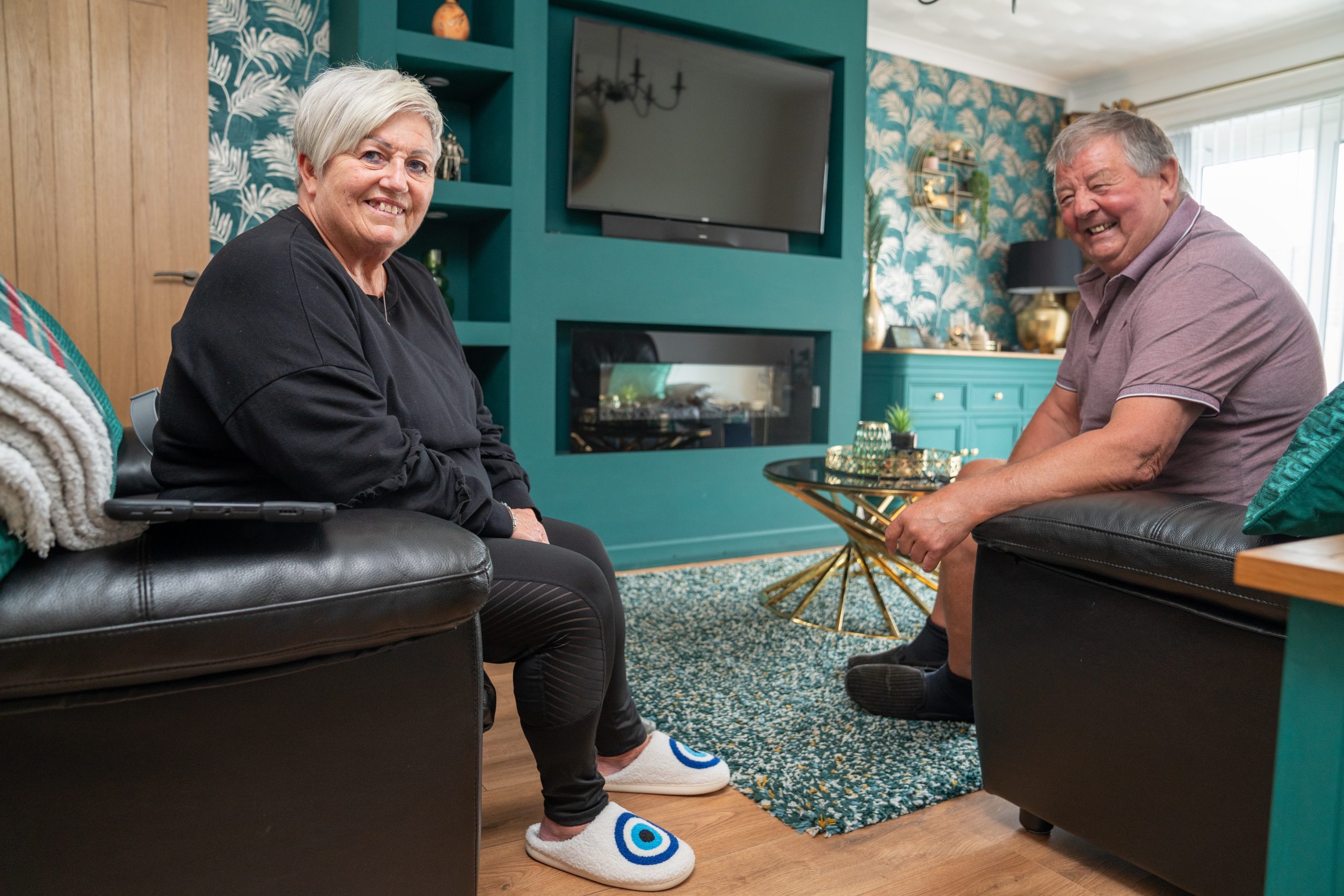 Trivallis Housing Landlord Wales An older woman and man sit in a living room with teal-painted walls and floral wallpaper. The woman, wearing a black outfit and slippers, and the man, in a purple shirt and black socks, smile at the camera. A TV and fireplace are mounted on the wall.