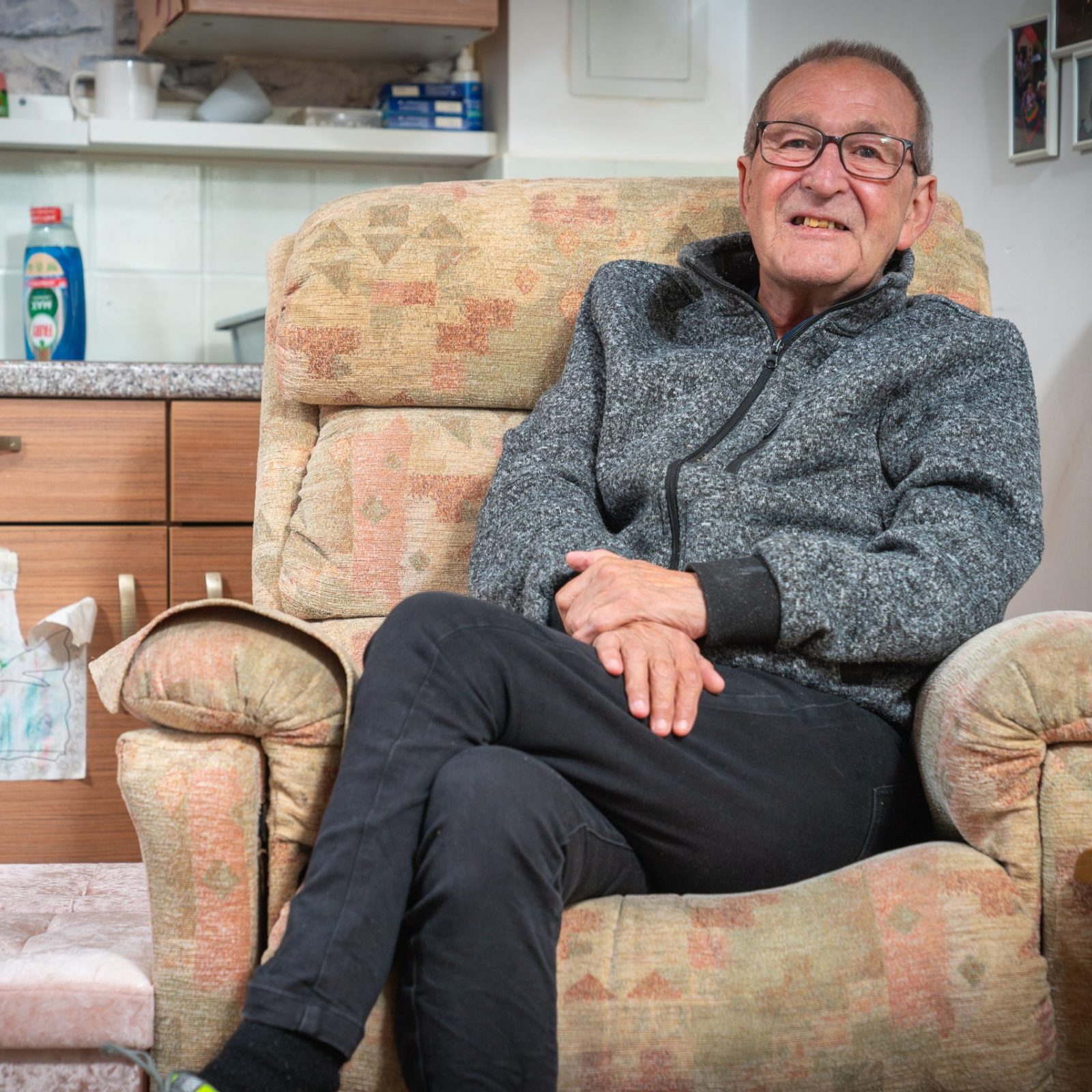 Trivallis Housing Landlord Wales An older man sits in a patterned armchair with his legs crossed. He wears glasses and a gray zip-up jacket. Behind him is a kitchen area with wooden cabinets and various household items. To his side is a small footstool.