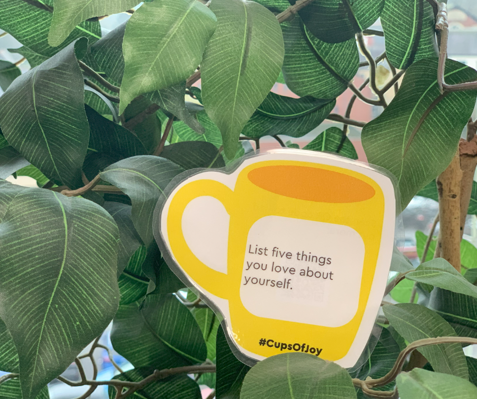 Trivallis Housing Landlord Wales A yellow coffee cup-shaped card is nestled among green leaves. The card reads "List five things you love about yourself." A hashtag, #CupsOfJoy, is printed below the message.