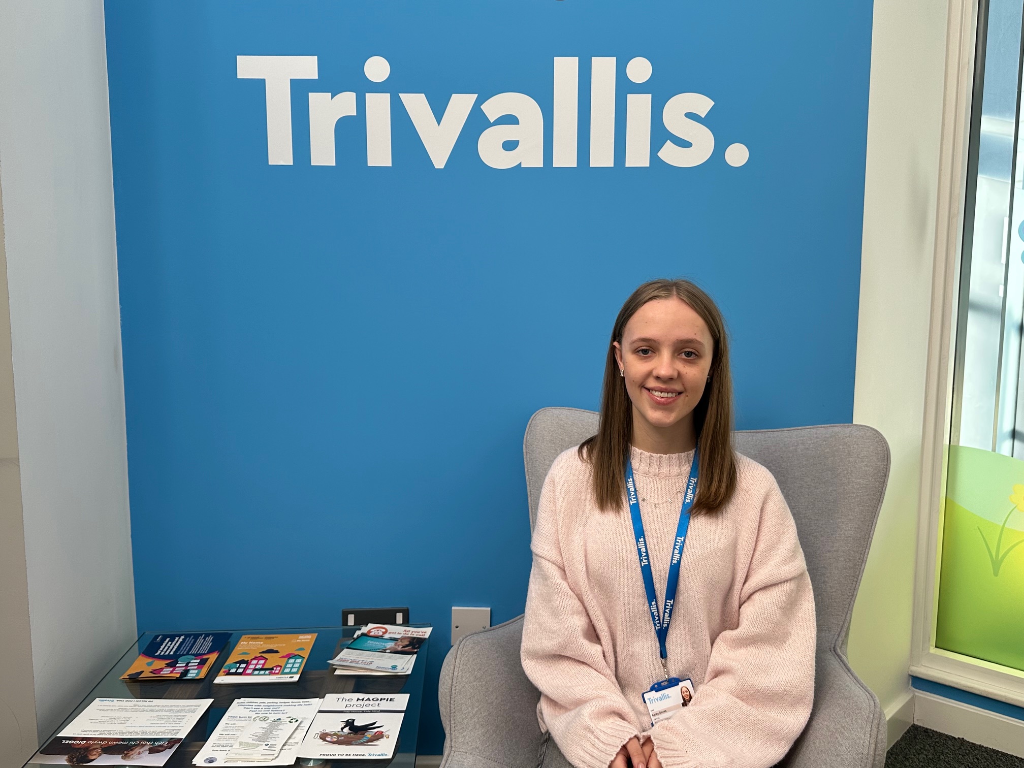 Trivallis Housing Landlord Wales A woman with long hair, wearing a light-colored sweater and a blue lanyard, sits in a gray chair. Behind her is a blue wall with the word "Trivallis." A table beside her has brochures and leaflets.