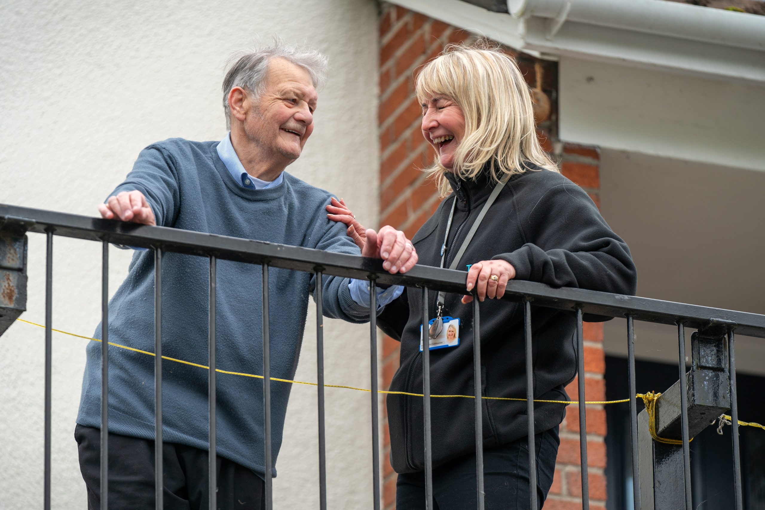 Trivallis Housing Landlord Wales An older man and a middle-aged woman stand smiling on a metal balcony railing. The man rests his arm on the rail, while the woman leans slightly forward. They appear to be engaged in a friendly conversation.