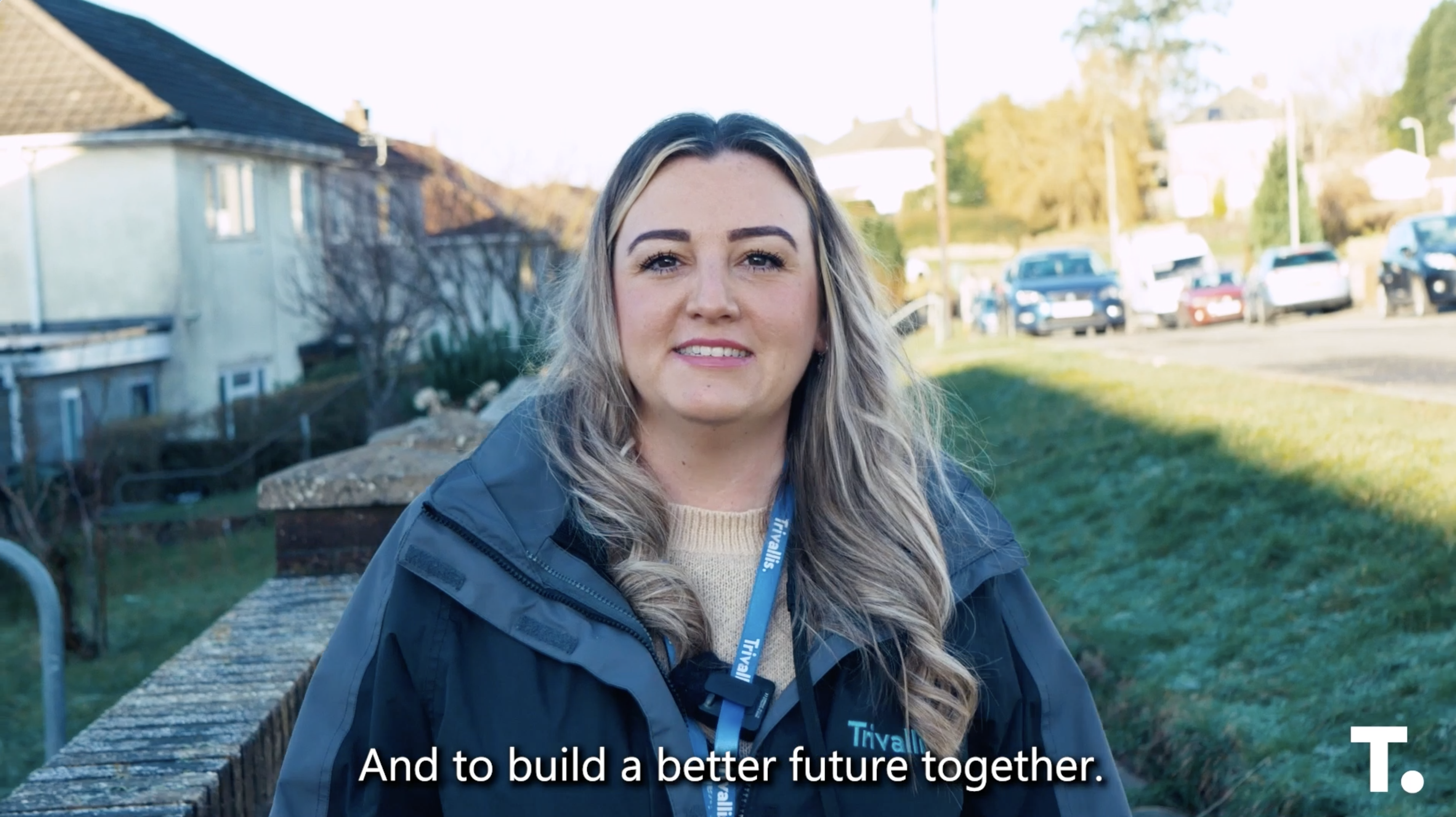 Trivallis Housing Landlord Wales A person with long hair, wearing a jacket, stands outdoors on a clear day. Houses and cars are visible in the background. The text reads, "And to build a better future together.