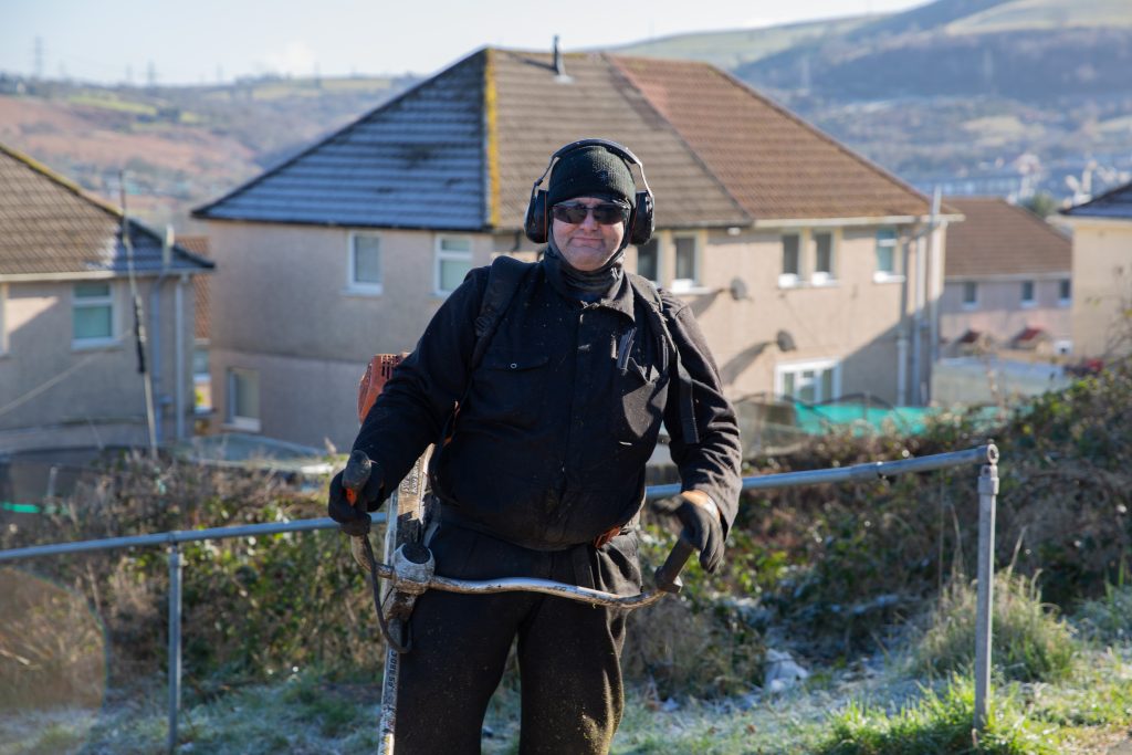 Trivallis Housing Landlord Wales A person in protective clothing and earmuffs operates a trimmer in a rural residential area. Houses and hills with a light dusting of frost are in the background. The person looks toward the camera.