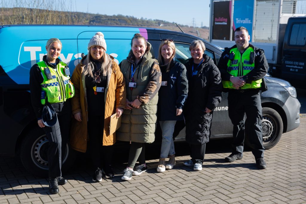 Trivallis Housing Landlord Wales A group of six people, including two police officers in uniform and four others in casual winter clothing, stand in front of a vehicle. They are outdoors on a paved area. The vehicle has the text "Trivallis" on it.