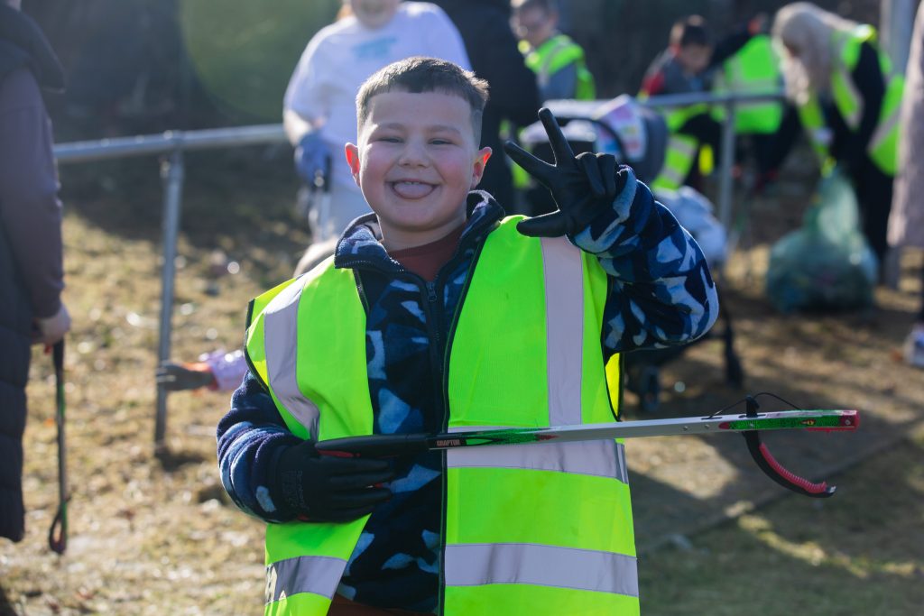 Trivallis Housing Landlord Wales A child in a yellow safety vest holds a litter picker and gives a peace sign outdoors. Other people in similar vests are in the background. It appears to be a group cleanup event in a grassy area.