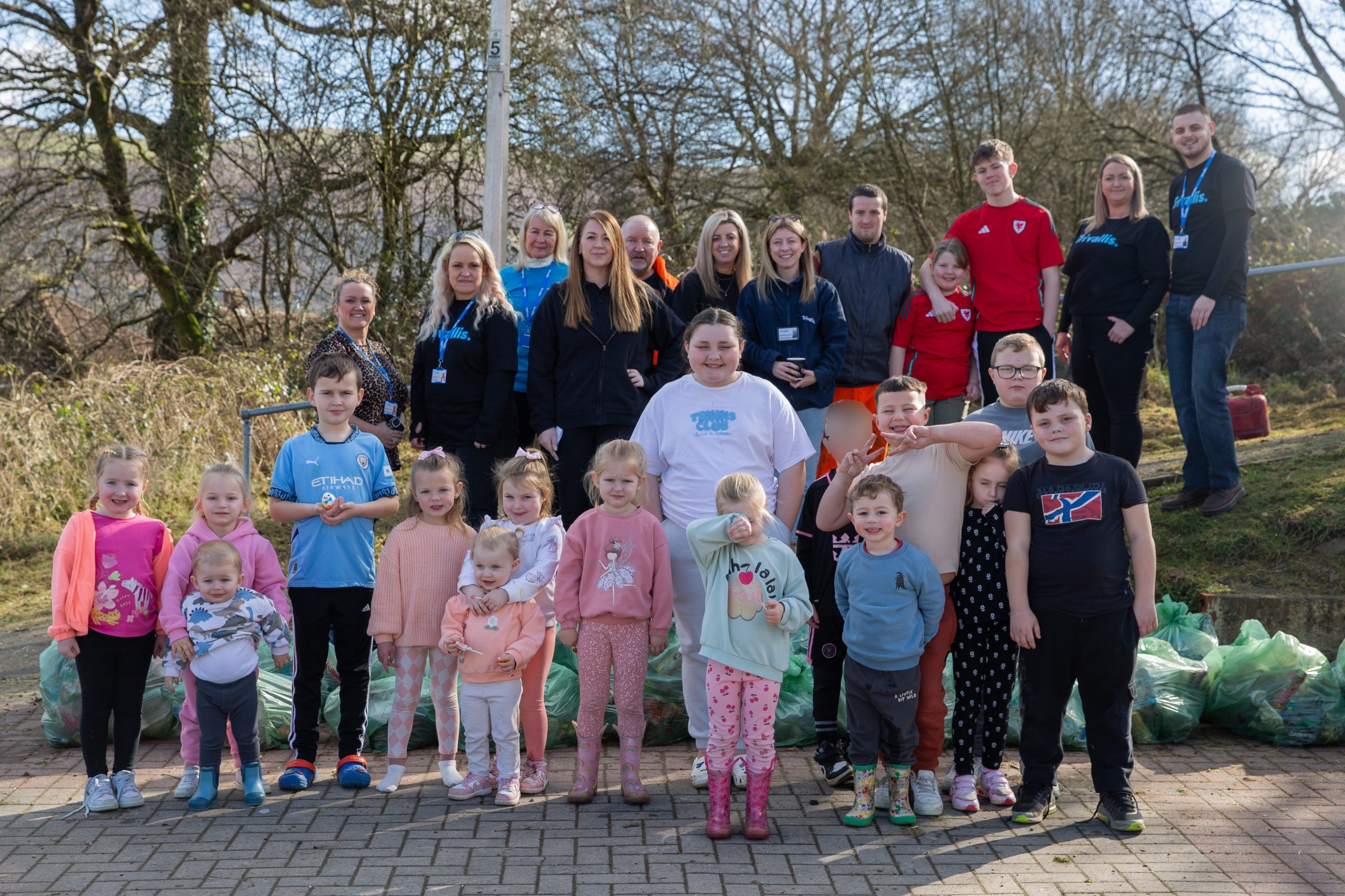 Trivallis Housing Landlord Wales A group of adults and children stand outdoors on a paved area. They are dressed in casual clothing and positioned in rows. A pile of green bags is visible in the background along with trees and a clear sky.