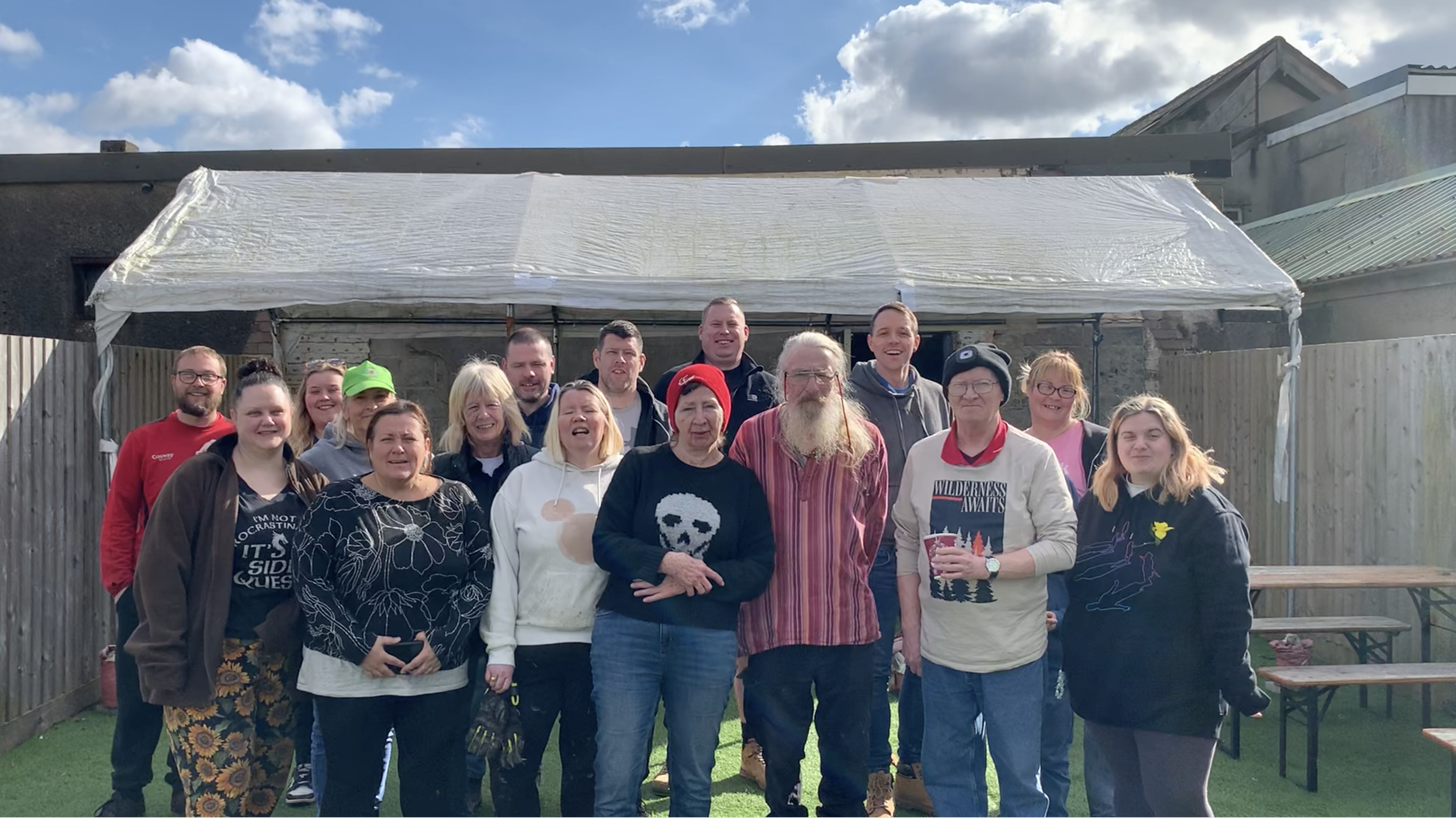 Trivallis Housing Landlord Wales A group of people standing together in a backyard setting. They are in front of a wooden building with a translucent roof. The sky is partly cloudy. Some are smiling, and they vary in clothing style and colors.