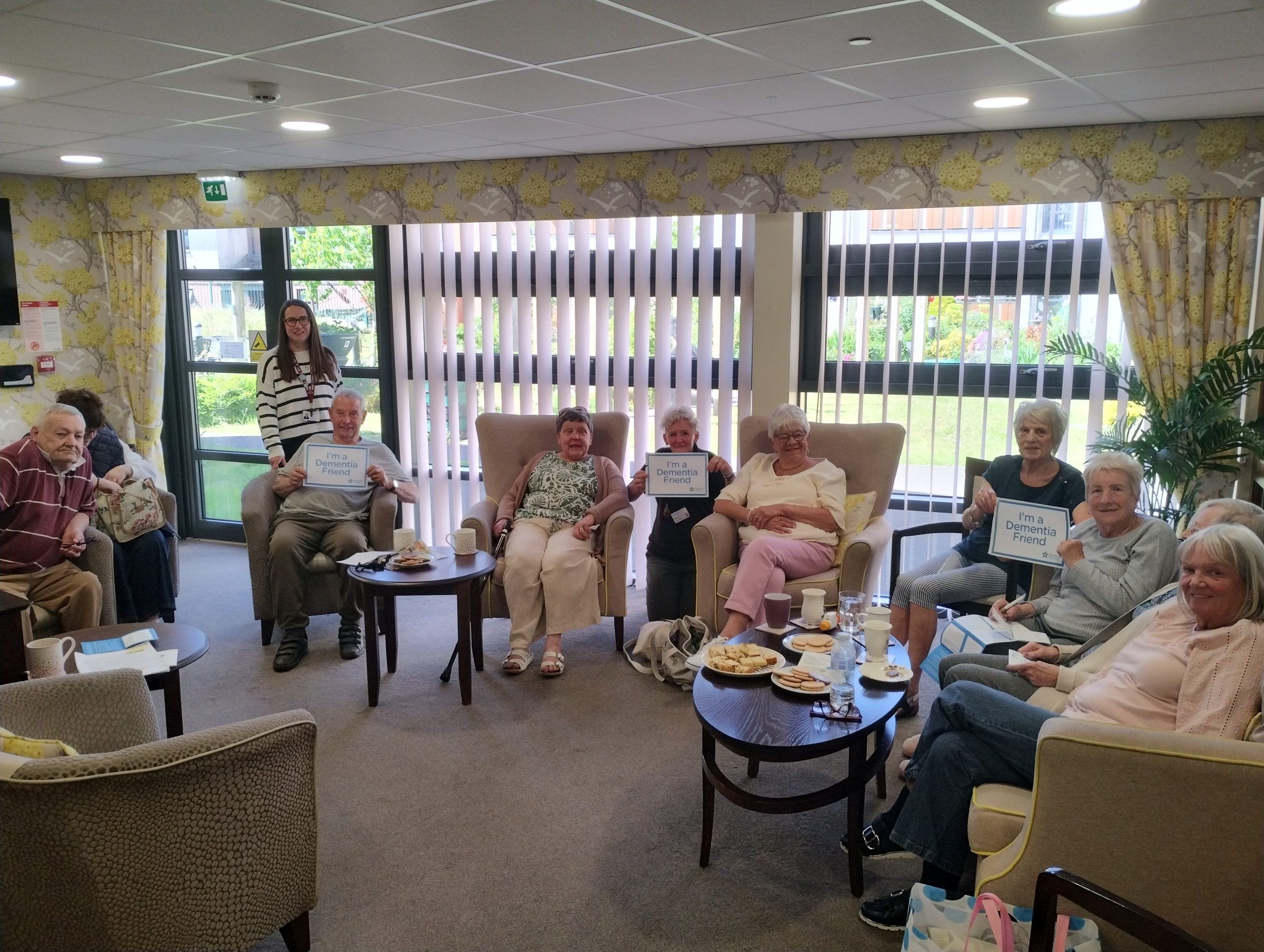 Trivallis Housing Landlord Wales A group of older adults sit in armchairs around small tables with snacks and drinks in a bright, communal room. Some people hold signs, and a younger woman stands behind the group, smiling. Large windows are in the background.