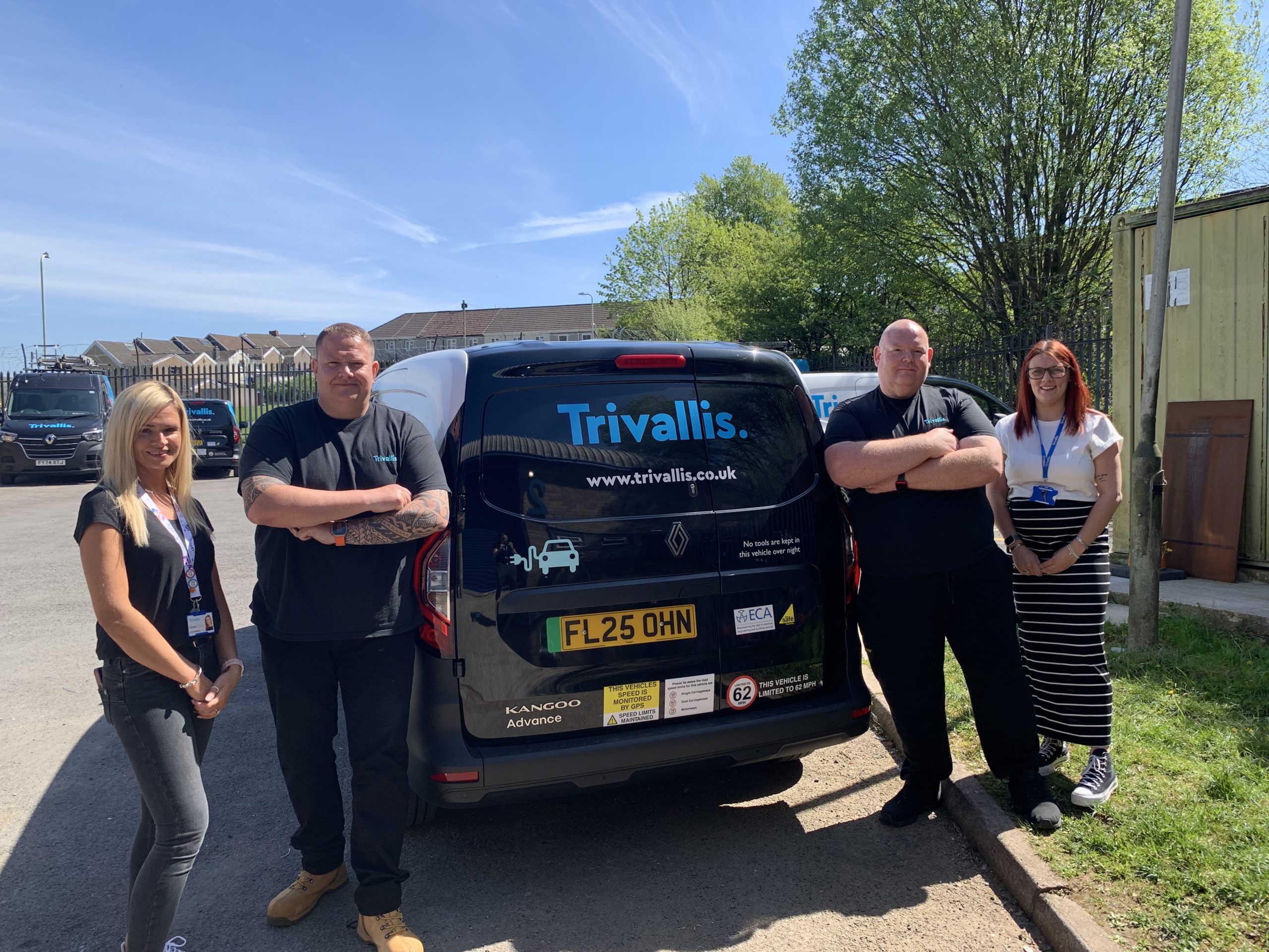 Trivallis Housing Landlord Wales Four people stand next to a black van with "Trivallis" branding and a website on the back. The van is parked outside on a sunny day beside a fence and trees. The group faces the camera and smiles.