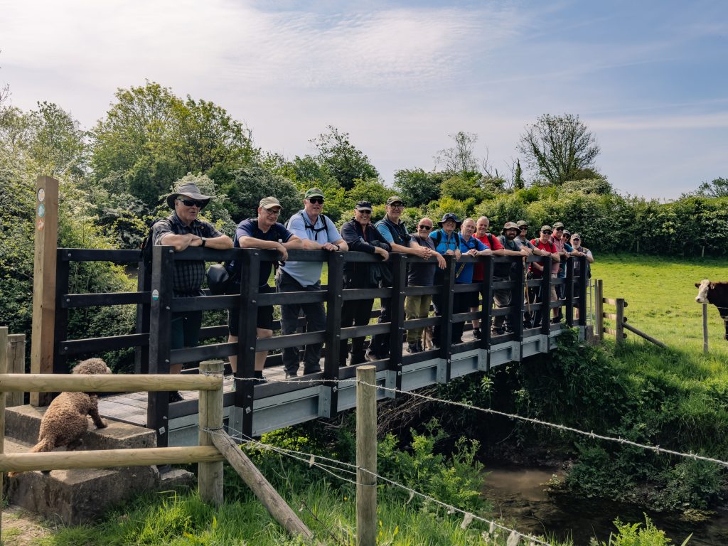 Trivallis Housing Landlord Wales A group of people stands side by side on a wooden bridge over a small stream, surrounded by trees and greenery on a sunny day. A dog is visible in the bottom left corner near the fence.