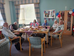 Trivallis Housing Landlord Wales Six elderly people are sitting around a table with food and drinks, decorated with Union Jack flags and bunting, in a brightly lit room. Red, white, and blue balloons and decorations are visible in the background.