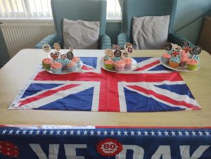 Trivallis Housing Landlord Wales A table with a Union Jack flag displays cupcakes with pink and yellow icing and various toppers. Two chairs are behind the table. A VE Day banner with "80 Years" is draped at the front of the table.