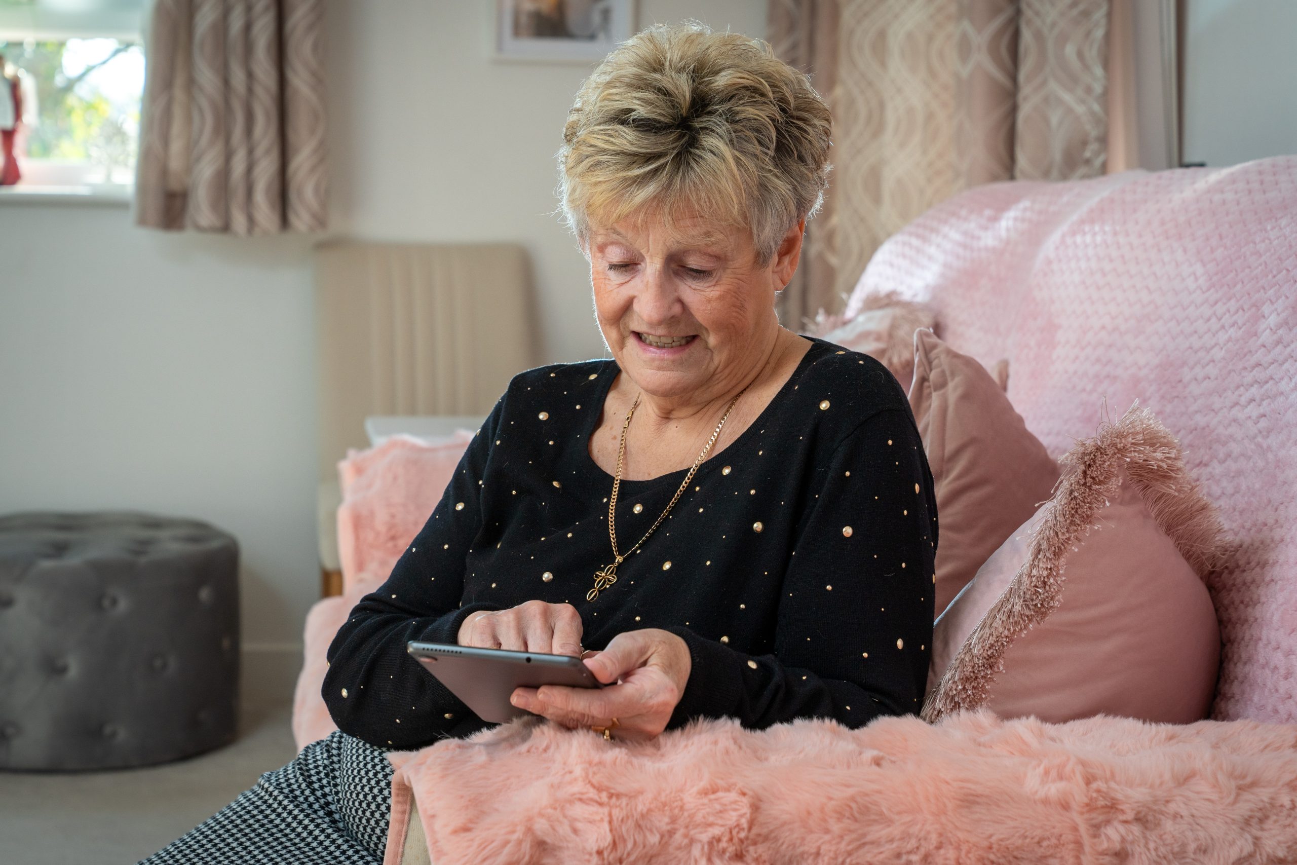 Trivallis Housing Landlord Wales An older woman sits on a pink sofa with textured cushions, using a tablet. She is wearing a black top with small gold dots and a gold necklace. The room is softly lit with neutral-colored curtains and decor.