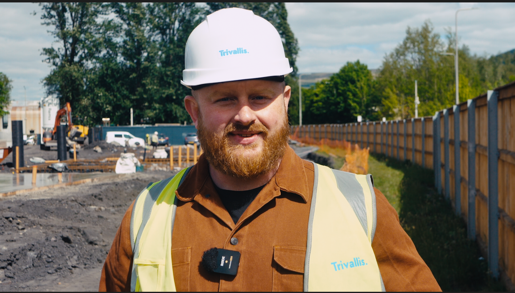 Trivallis Housing Landlord Wales A man with a beard wearing a white hard hat and yellow safety vest stands at a construction site. He has a microphone clipped to his shirt and construction work is visible in the background.