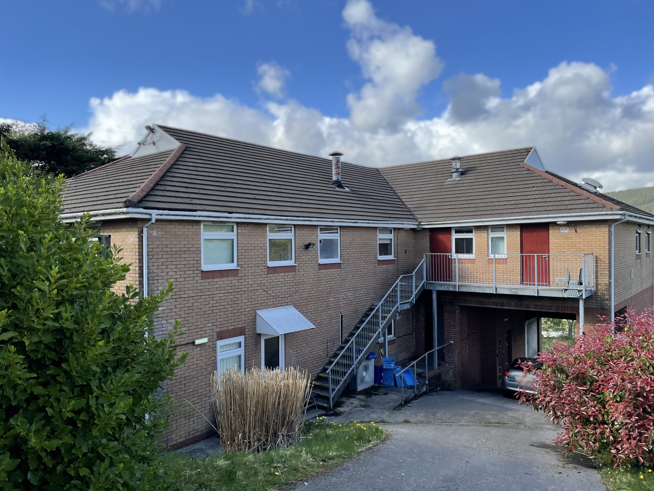 Trivallis Housing Landlord Wales A two-story brick building with an external metal staircase leading to an upper walkway. There is a driveway below the walkway, a parked car, and shrubs in the foreground. The sky is partly cloudy.