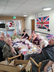 Trivallis Housing Landlord Wales A group of elderly people sit around a table eating food, decorated with Union Jack flags and VE Day signs, in a communal room with beige walls and a bulletin board in the background.