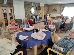 Trivallis Housing Landlord Wales A group of older women sit around a table with food and drinks in a lounge, holding British-themed props. Some women are smiling, and decorations are visible in the background.