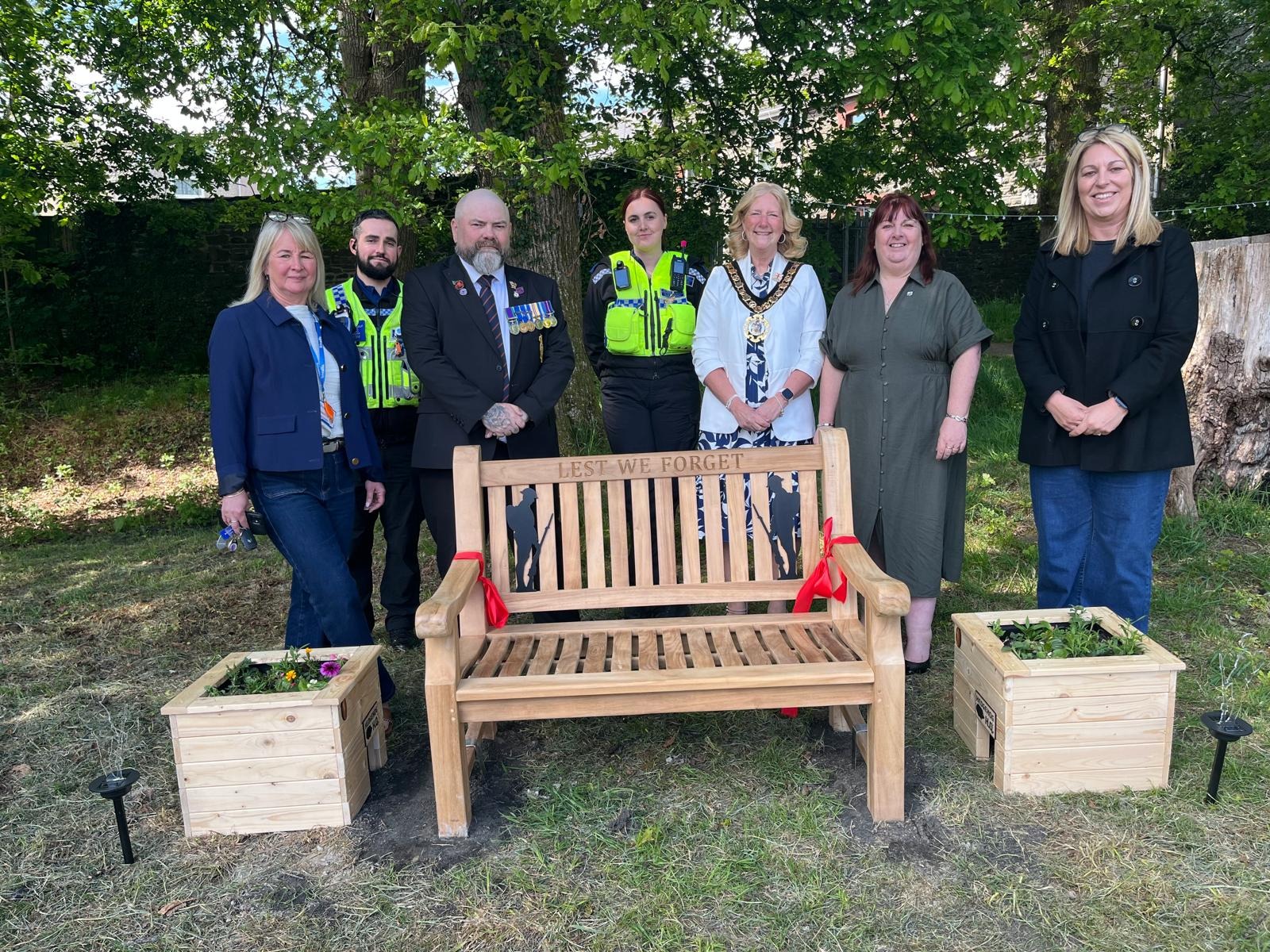 Trivallis Housing Landlord Wales Seven people stand outdoors behind a wooden bench engraved with "LEST WE FORGET," flanked by two wooden planters with flowers; two people are in police uniforms, and trees are visible in the background.