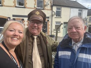 Trivallis Housing Landlord Wales Three adults stand outdoors smiling at the camera. One woman is on the left, a man in a brown military-style uniform and glasses is in the middle, and another man in a blue jacket is on the right. Houses are visible in the background.