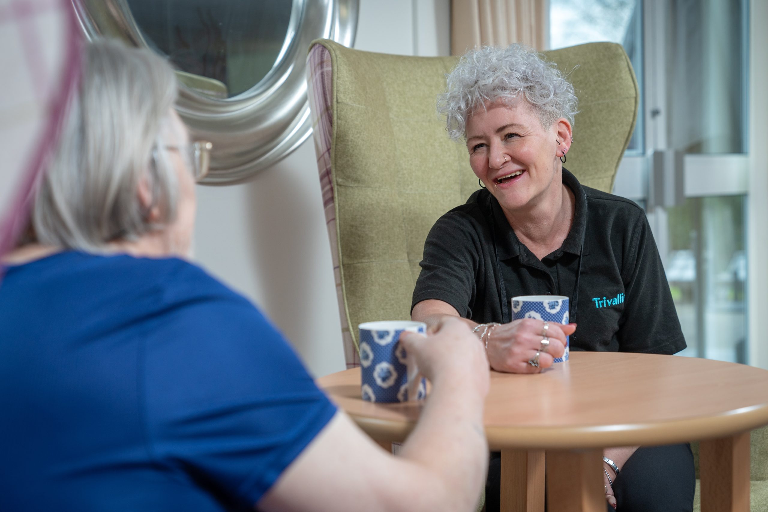 Trivallis Housing Landlord Wales Two women sit across from each other at a small wooden table, holding patterned mugs. One woman wears a blue shirt, while the other, wearing a black polo shirt, smiles and leans forward in her chair.