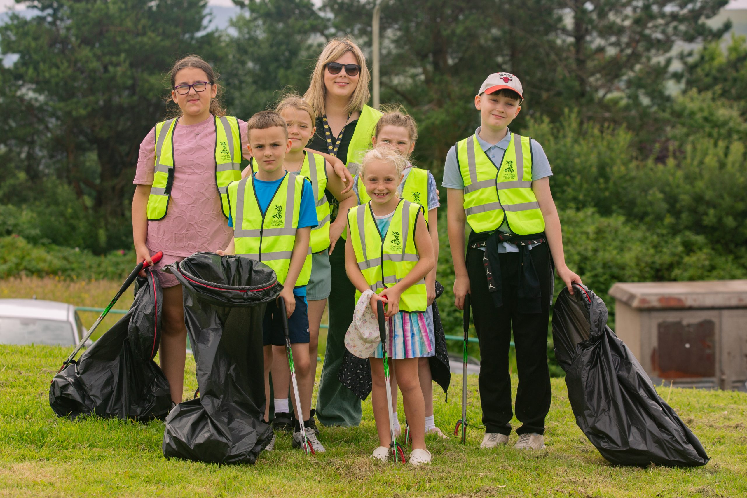 Trivallis Housing Landlord Wales A group of children and an adult, all wearing yellow safety vests, stand on grass holding litter-picking tools and black garbage bags, with trees and parked cars in the background.