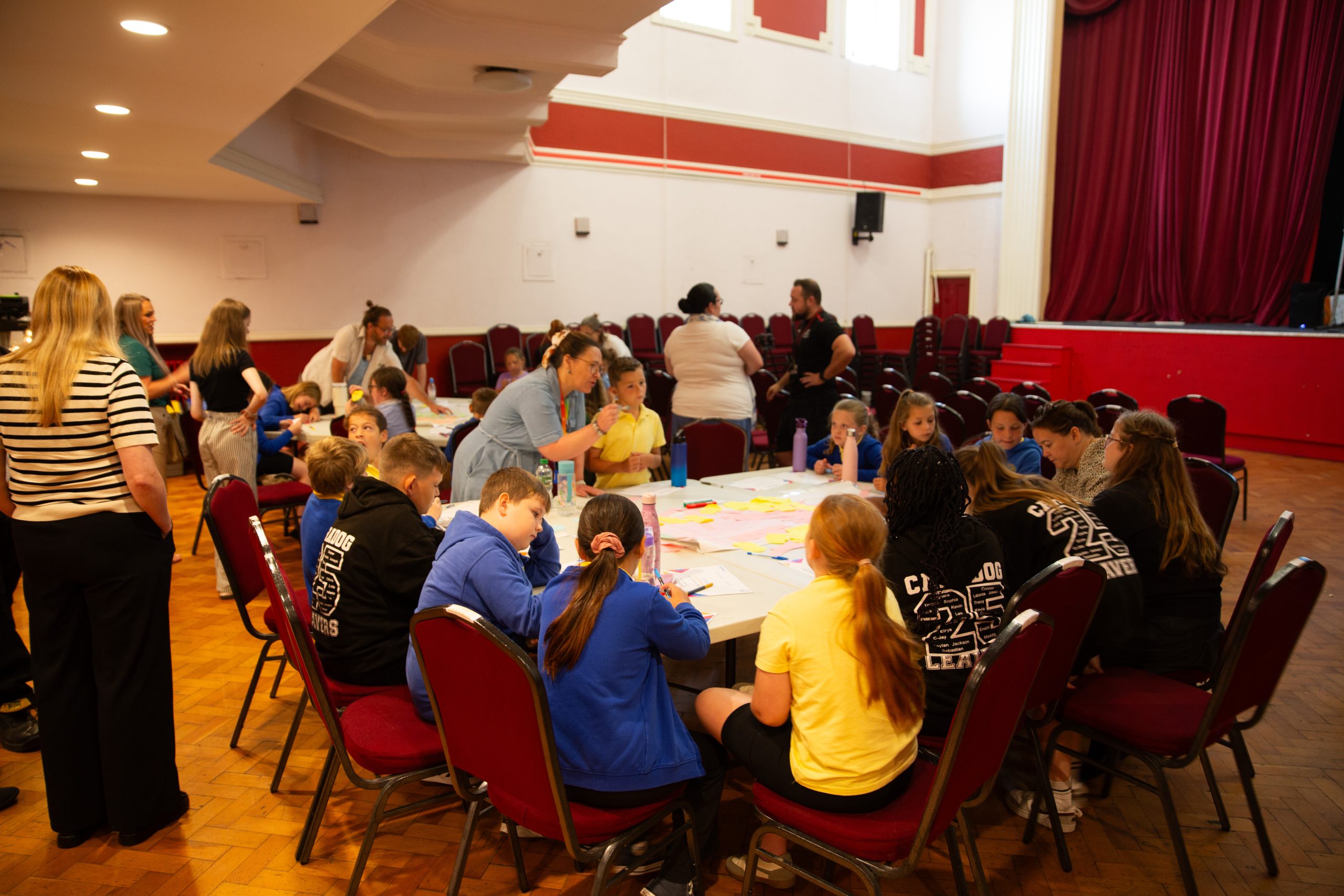 Trivallis Housing Landlord Wales A group of children and adults are gathered around tables in a large room, working on activities with colorful papers and markers. Some adults stand nearby, while others sit with the children. The room has red chairs and a stage.