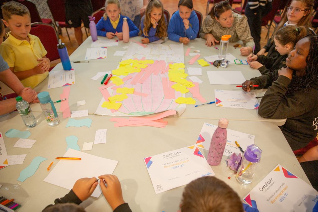 Trivallis Housing Landlord Wales Children sit around a table covered with papers, markers, certificates, water bottles, and cut-out shapes, engaging in a group activity in a brightly lit indoor setting.