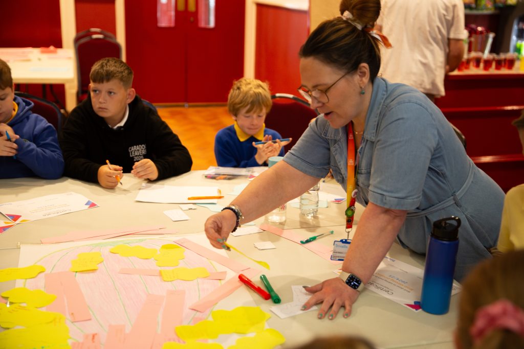 Trivallis Housing Landlord Wales A woman leans over a table covered with paper and yellow sticky notes, engaging with children seated around her. The kids appear focused on writing or drawing. Markers and papers are scattered on the table.