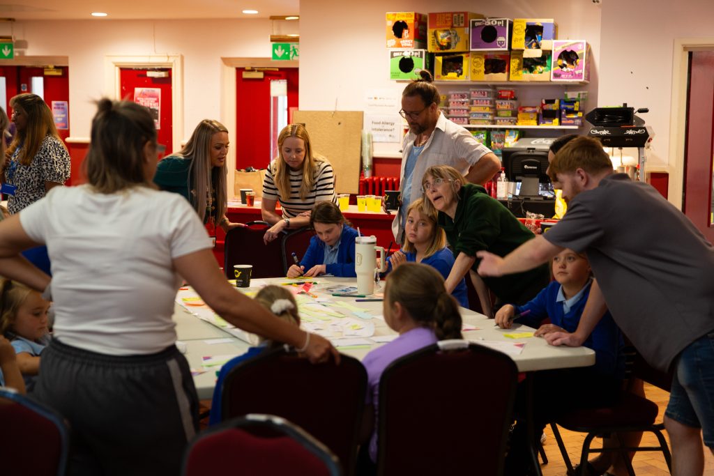 Trivallis Housing Landlord Wales Several adults and children are gathered around a table covered with papers and cups in a brightly lit room. The adults appear to be guiding or assisting the children, who are seated and wearing blue uniforms.