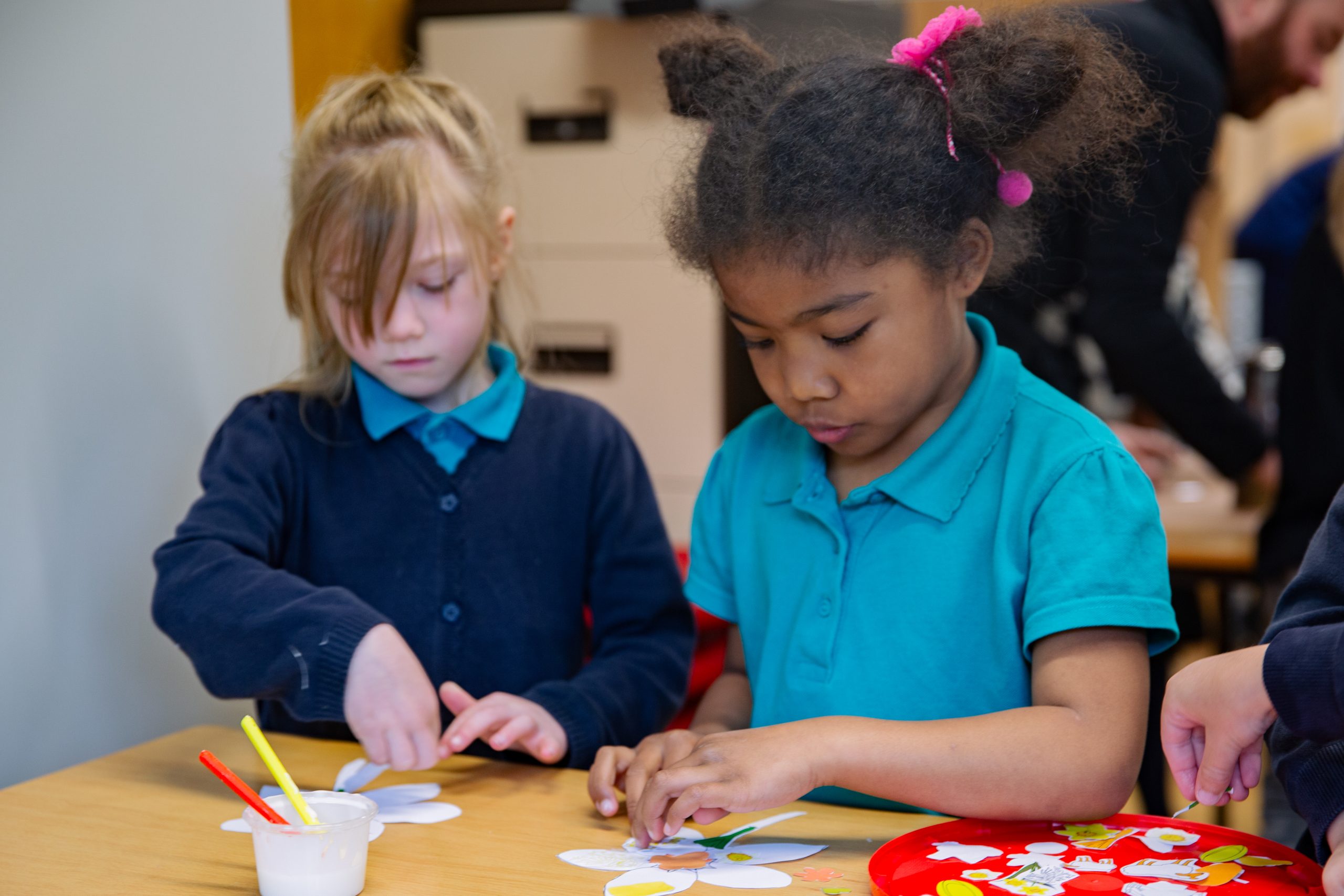 Trivallis Housing Landlord Wales Two young girls sit at a table in a classroom, focused on making paper crafts with glue and cut-out shapes. Cups with paintbrushes and a tray with paper pieces are on the table.
