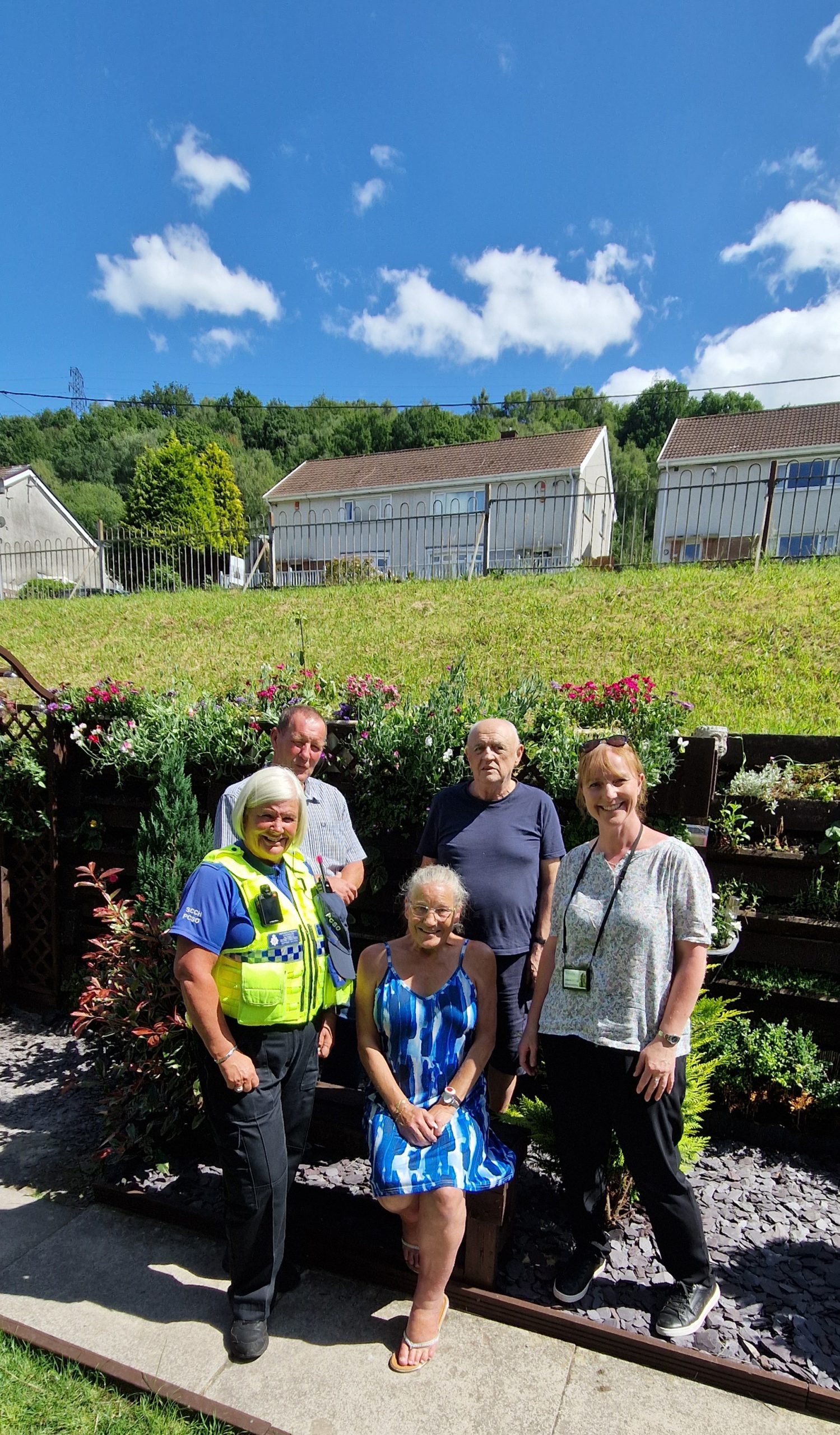 Trivallis Housing Landlord Wales Five adults, including a uniformed police officer and four casually dressed individuals, stand and sit in front of a flower bed in a garden, with houses and greenery visible in the background under a blue sky.
