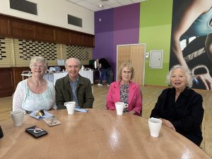 Trivallis Housing Landlord Wales Four older adults sit at a round wooden table with white mugs and plates. Behind them, a person stands at a buffet table. The room has colorful green and purple walls and large mural artwork.